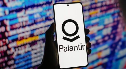 A hand holding a mobile phone displaying the Palantir logo, in front of a screen showing computer code